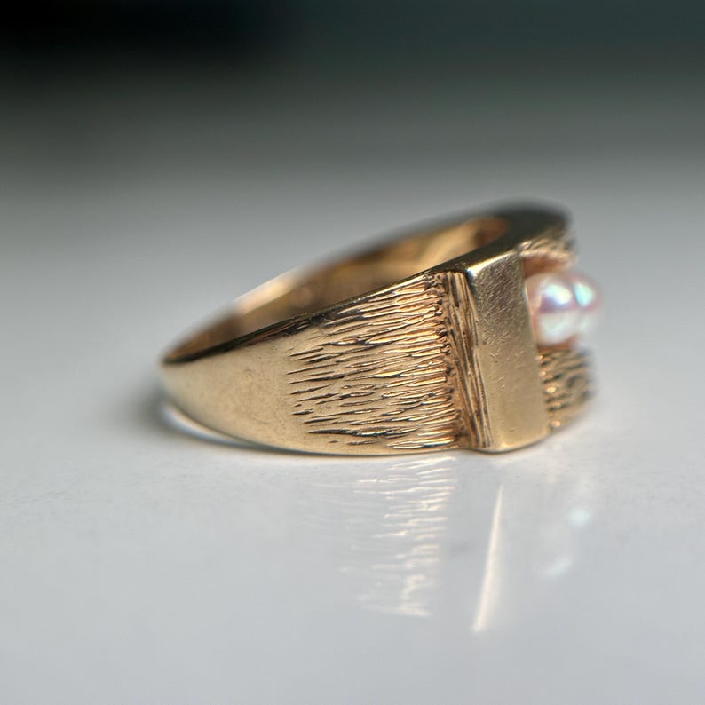 May include: A gold-colored ring with a textured band and a rectangular setting. The setting holds a small, iridescent pearl. The ring has a classic, vintage design.