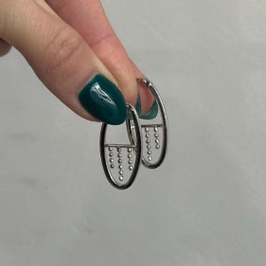 May include: Silver hoop earrings with a delicate chain of small beads dangling from the bottom. The earrings are oval shaped and have a minimalist design.