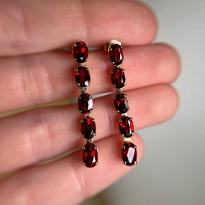 May include: A pair of gold-coloured drop earrings, each with five oval-cut, deep red gemstones. The gemstones are evenly spaced along the length of the earrings, creating a linear design. The earrings are displayed against a blurred background.