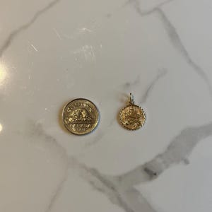 Puede incluir: Un colgante dorado con un peque&ntilde;o lazo para una cadena junto a una moneda canadiense. El colgante es redondo con un dise&ntilde;o detallado. La moneda tambi&eacute;n es dorada y tiene texto y un dise&ntilde;o.