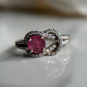 May include: A silver ring with a pink gemstone set in a knot design. The ring is surrounded by smaller clear gemstones.