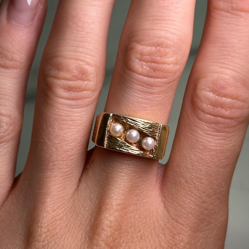 May include: A gold-colored ring featuring three small, round pearls set in a textured rectangular design. The ring has a classic, elegant style.