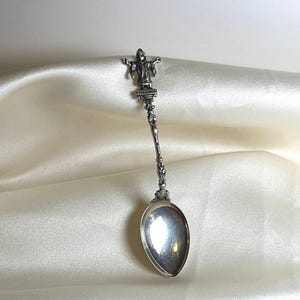 May include: A silver spoon with an ornate handle and a figure at the top. The spoon features a polished bowl and detailed design. The figure is of a person with outstretched arms. This spoon is likely a collectible or decorative item.