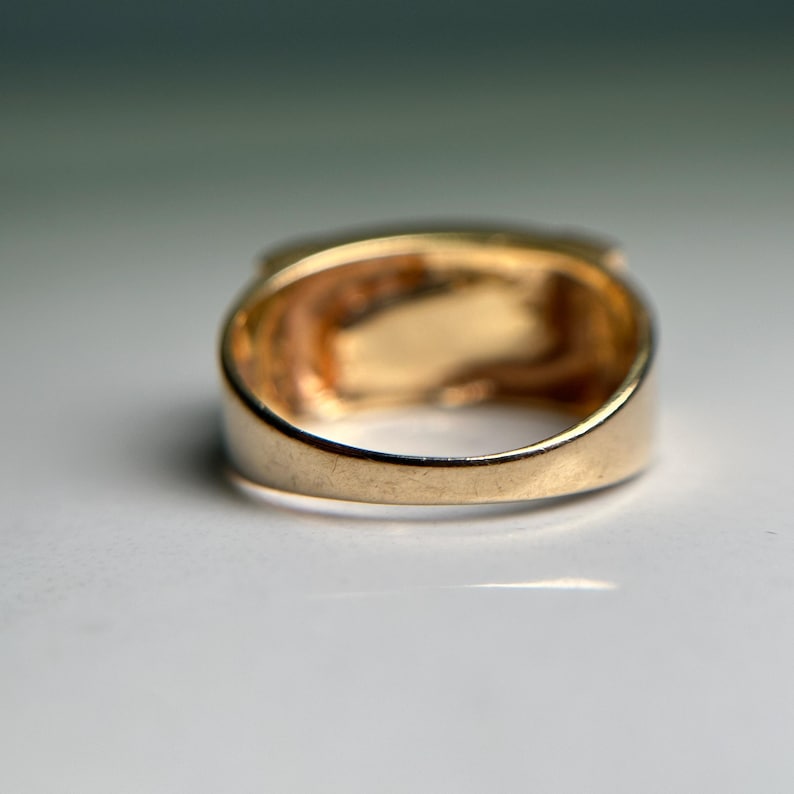 May include: A close-up shot of a gold ring. The ring has a rectangular face with a detailed interior design. The band is thick and rounded, and the ring appears to be made of solid gold.