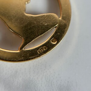May include: Close-up of a gold-colored pendant with a cut-out design. The pendant has the number "750" stamped on the back.