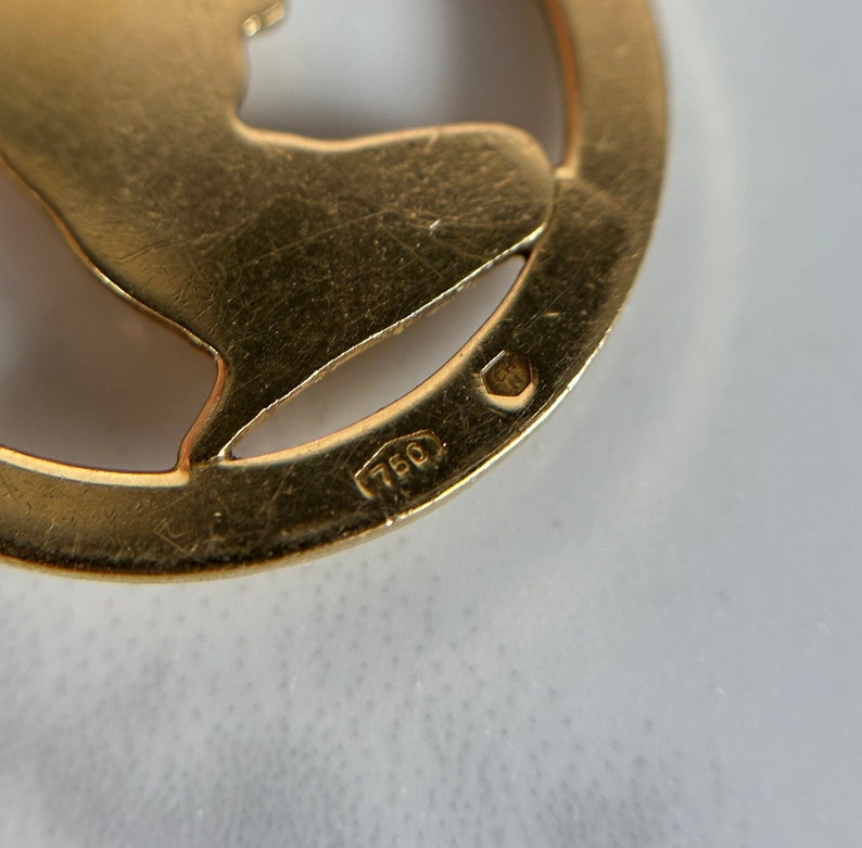May include: Close-up of a gold-colored pendant with a cut-out design. The pendant has the number "750" stamped on the back.