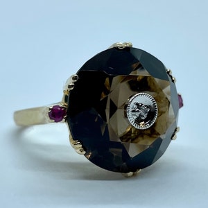 May include: A gold ring featuring a large, faceted, dark brown gemstone surrounded by smaller, red gemstones. A small, white diamond is set in the centre of the gemstone.