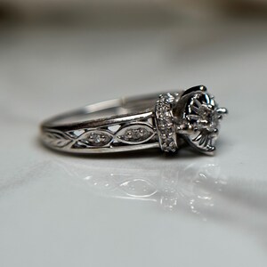 May include: A silver ring with a large, round, prong-set diamond in the center. The band is intricately designed with a delicate, woven pattern and smaller diamonds.