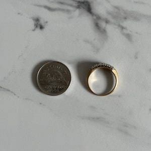 May include: A gold band ring with a row of small, clear stones set in the band. The ring is next to a Canadian 25-cent coin dated 2008.