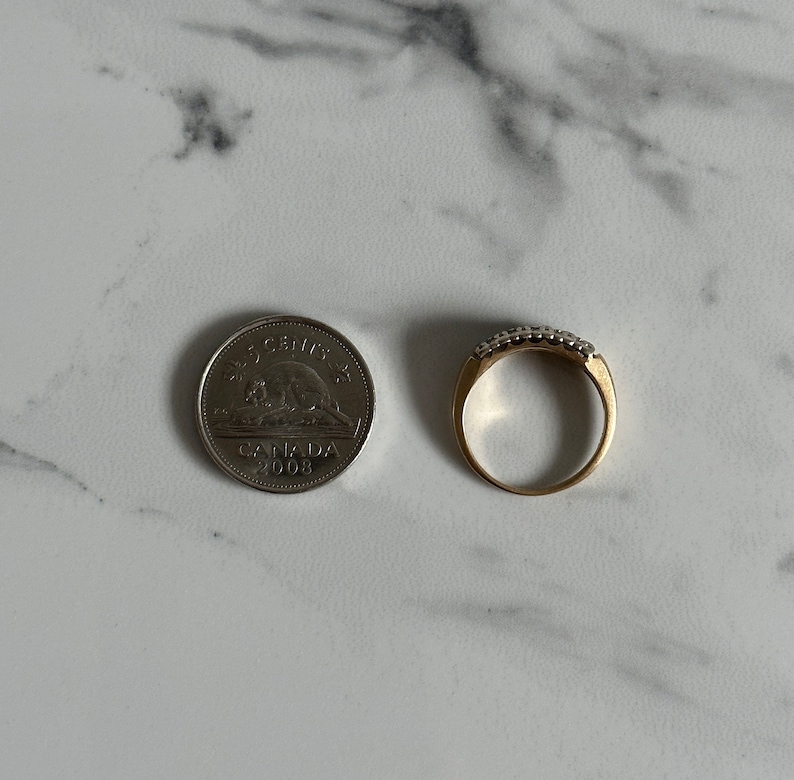 May include: A gold band ring with a row of small, clear stones set in the band. The ring is next to a Canadian 25-cent coin dated 2008.