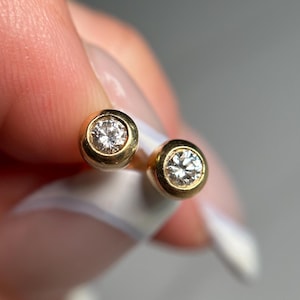 May include: A pair of gold stud earrings with a bezel setting. Each earring features a round, clear diamond. The earrings are displayed against a soft, neutral background, highlighting their sparkle and design.