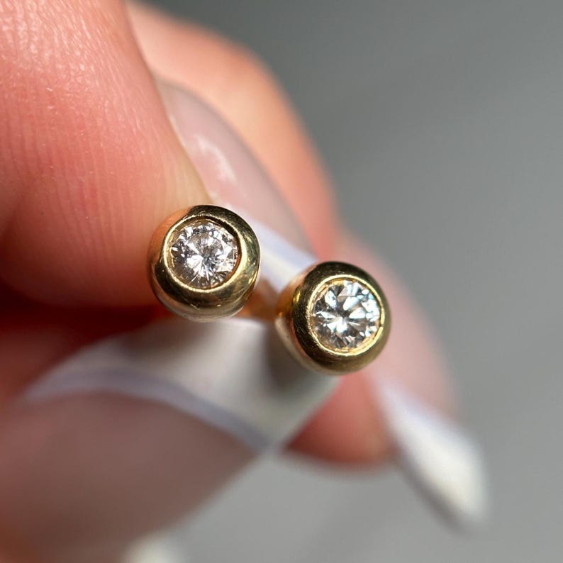 May include: A pair of gold stud earrings with a bezel setting. Each earring features a round, clear diamond. The earrings are displayed against a soft, neutral background, highlighting their sparkle and design.