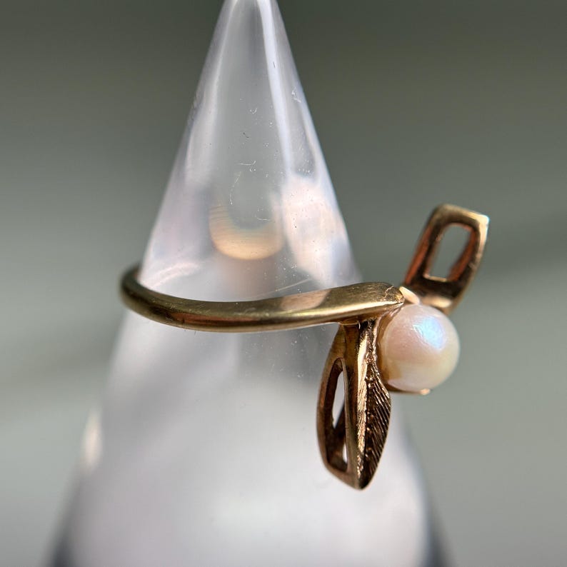 May include: A gold-colored ring with a pearl accent. The ring features a bow-like design with a small, round pearl at the center. The ring is displayed on a clear, cone-shaped stand against a soft gray background.