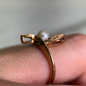 May include: A gold-toned ring featuring a small, round pearl at the center. The ring has a unique design with two openwork, leaf-like shapes on either side of the pearl. The band is smooth and simple, creating an elegant and classic look.
