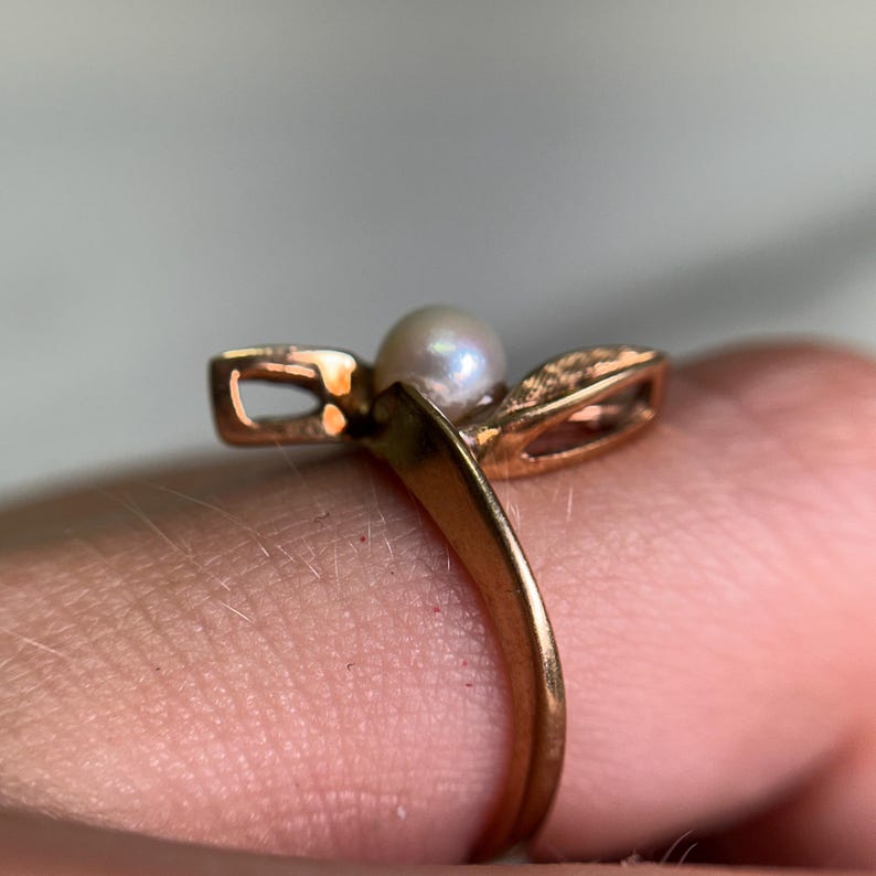 May include: A gold-toned ring featuring a small, round pearl at the center. The ring has a unique design with two openwork, leaf-like shapes on either side of the pearl. The band is smooth and simple, creating an elegant and classic look.