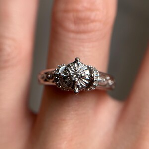 May include: A silver engagement ring with a large, round, clear diamond set in a six-prong setting. The band is decorated with intricate details.