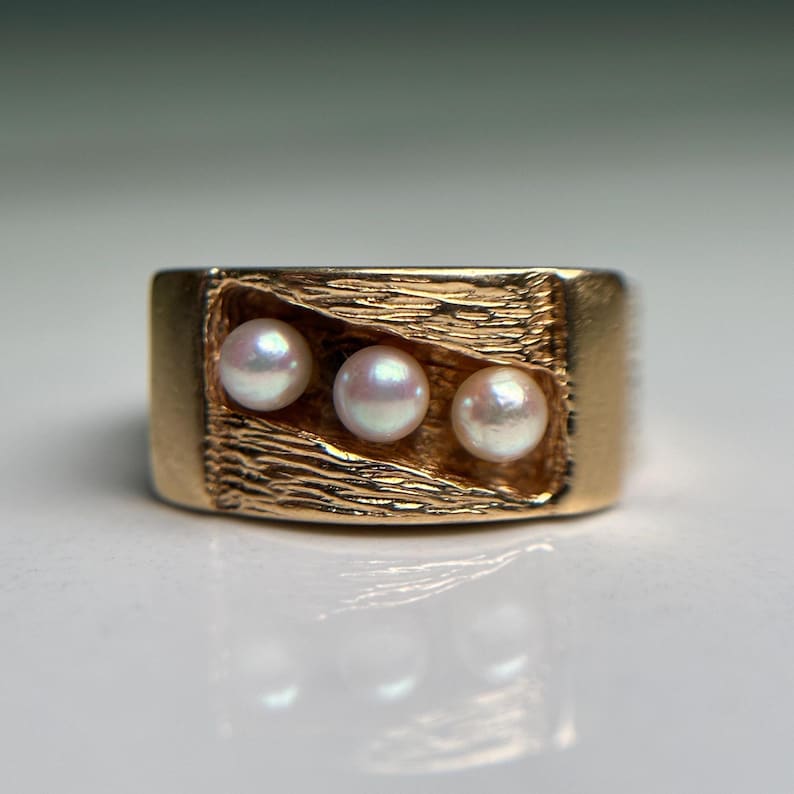 May include: A gold-colored ring featuring three small, round pearls set in a textured, rectangular design. The ring has a thick band and a polished finish, reflecting light.