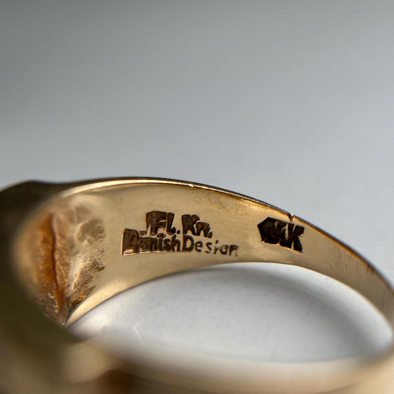 May include: Close-up of a gold ring with the text "JFL. Kr. Danish Design" and "14K" engraved on the inside. The ring has a smooth, polished surface and a classic design.