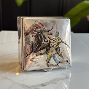 May include: A silver box with a detailed engraving of a bullfight scene. The artwork depicts a bull charging a matador, with a red cape and mountain backdrop. The box is square and has a hinged lid.