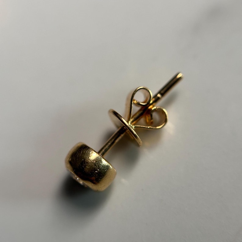 May include: A single gold stud earring with a round, bezel-set gemstone. The earring post and butterfly backing are also gold. The earring is set against a neutral background, highlighting its simple elegance.