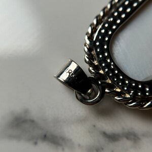 May include: Close-up of a silver pendant with a textured, rope-like border. The pendant is shaped like a U and has a small, engraved marking on the back that reads "925".