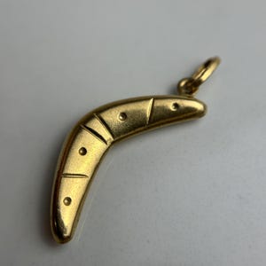 May include: A gold-toned boomerang-shaped pendant featuring a smooth, curved surface with engraved lines and small circular indentations along its length. The pendant includes a small circular jump ring attached to one end for hanging purposes.