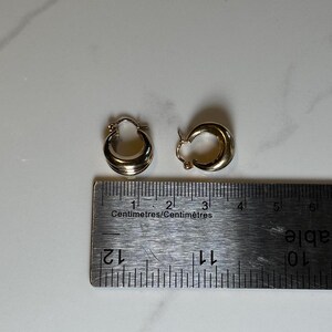 May include: A pair of gold hoop earrings measuring approximately 1 cm in diameter. The earrings are shown next to a ruler for size reference.