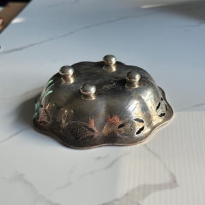 May include: A silver bowl with a scalloped edge and floral cutouts. The bowl has four feet and is made of metal.