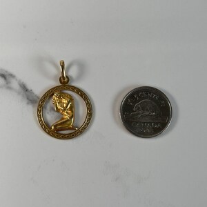 May include: Gold pendant featuring a detailed depiction of a woman kneeling, framed by a circular design with a textured border.