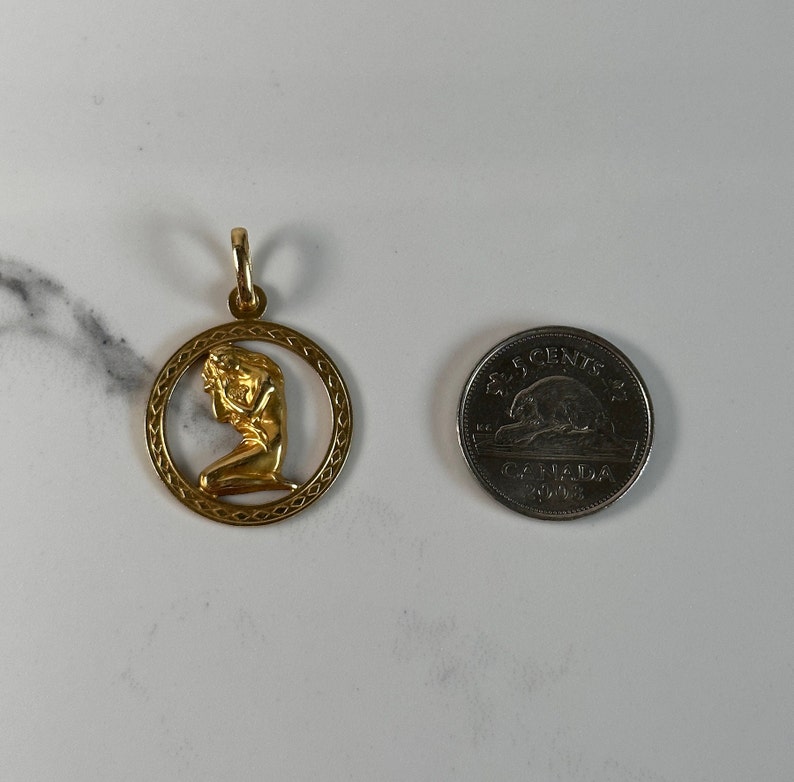 May include: Gold pendant featuring a detailed depiction of a woman kneeling, framed by a circular design with a textured border.