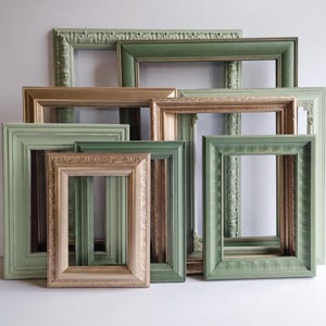 May include: A collection of rectangular picture frames in various sizes, featuring sage green and metallic gold finishes. The frames display diverse textures, including smooth surfaces, ornate carved detailing, and stepped edges, arranged in a layered stack.