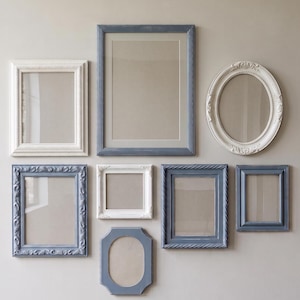 May include: A gallery wall arrangement featuring seven empty picture frames in various sizes and shapes. The collection includes rectangular and oval frames finished in shades of matte white and muted slate blue, with ornate and smooth border details.