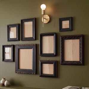May include: A gallery wall arrangement featuring eight black picture frames of varying sizes and styles, including smooth and ornate borders, surrounding a single brass wall sconce with a round base and a glowing white spherical light bulb.