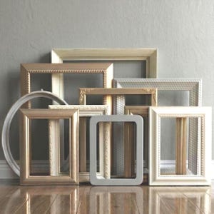 May include: A collection of empty picture frames in various shapes and sizes, including rectangular and oval designs. The frames feature diverse finishes such as metallic gold, matte cream, soft white, and light gray, with ornate and smooth borders.