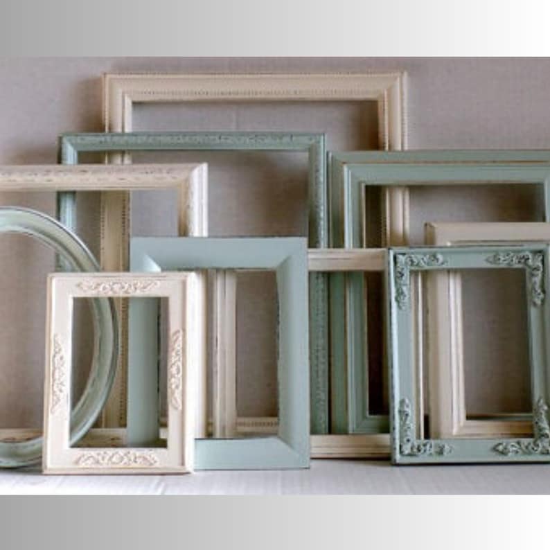 A gallery wall collection of assorted vintage picture frames painted jade green and white. Frames are distressed. Many frames have ornate details while others are more simple styles. Picture frames are various sizes, shapes and styles. The frames have a distressed, antique look.