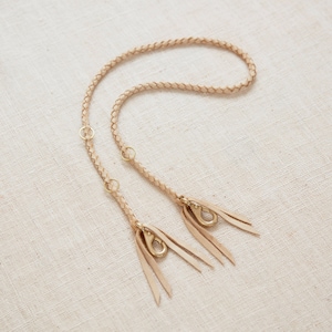 May include: A light brown leather strap with two brass clasps and tassels. The strap is braided and has a decorative design.