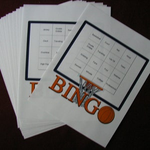 May include: A stack of white basketball bingo cards with a black and orange design. Each card features a bingo grid with basketball-related terms and the word "BINGO" in orange. An orange basketball graphic is also included.