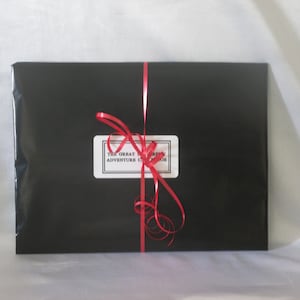 May include: A rectangular package wrapped in black paper, tied with a red ribbon. A white label with black text reads "THE GREAT ADVENTURE CLUB". The ribbon is tied in a bow and curls. The package is on a white surface.