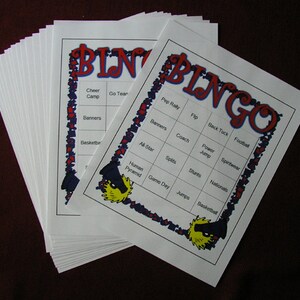 May include: A stack of white Bingo cards with red and blue decorative borders. The word "BINGO" is in large red letters at the top. Each card has a grid of squares with words like "Cheer Camp", "Banners", and "Basketball".