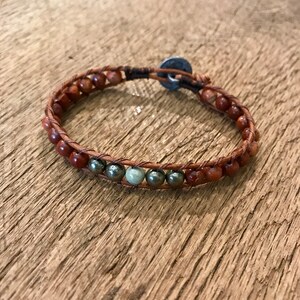 Könnte beinhalten: Ein braunes Lederarmband mit roten und goldenen Perlen. Das Armband hat einen silbernen Verschluss.