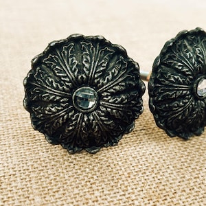 May include: Two decorative curtain tiebacks. Each tieback features a dark, ornate, floral design with a clear, round, crystal-like center. The tiebacks are made of a dark metal and are set against a neutral, textured background.