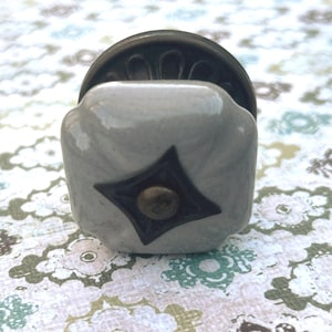 May include: A square, light gray cabinet knob with a dark bronze diamond design and a bronze screw. The knob has a dark bronze circular base. The background is a floral pattern in shades of green, brown, and blue.