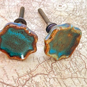 May include: Two ceramic cabinet knobs with a unique, organic shape. The knobs feature a teal and brown color scheme with a speckled texture. Each knob has a threaded metal post for easy installation. These decorative knobs add a touch of artistic flair to any furniture.
