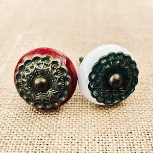 May include: Two decorative cabinet knobs. One is red with a bronze-colored floral design, and the other is white with a similar design. The knobs have a round shape and are likely made of ceramic or porcelain.