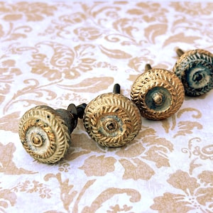May include: Four antique-style cabinet knobs with a distressed gold finish. Each knob has a round, textured design with a central circular detail. The knobs are arranged in a row against a floral patterned background.