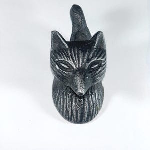 May include: A dark gray fox-shaped bottle stopper. The fox has detailed fur texture and pointed ears, with a tail extending upwards. The stopper is made of a solid material and sits upright on a white surface.