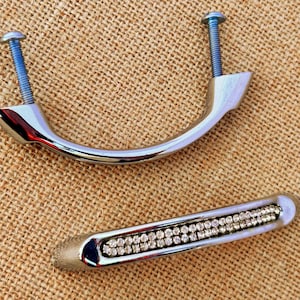 May include: Two silver cabinet pulls with screws. One is a curved handle, the other is a rectangular handle with a row of small, clear rhinestones. Both are on a textured, tan surface.