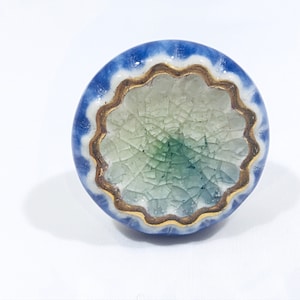 May include: A round ceramic cabinet knob with a blue and white outer edge, a gold scalloped inner edge, and a crackled green and white center. This decorative knob is suitable for furniture and home decor projects.