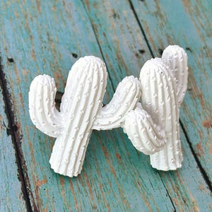 May include: Two white cactus-shaped cabinet knobs. The knobs are made of a textured material and have a screw on the back for installation. The knobs are set against a weathered, turquoise-colored wooden surface.