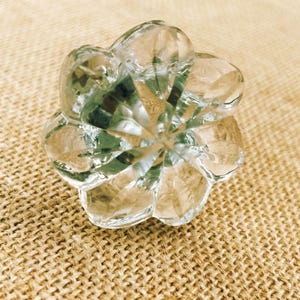 May include: A clear, faceted glass knob with a flower-like design. The knob has a central point with radiating petals, reflecting light. The background is a textured, tan fabric.